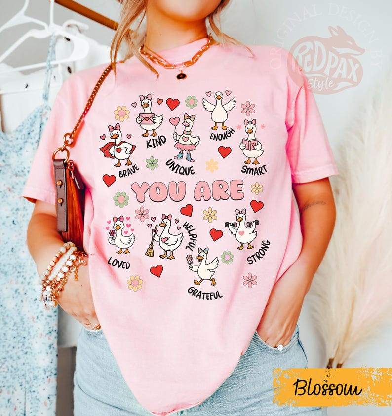 May include: A pink t-shirt with the words "YOU ARE" and illustrations of geese with positive affirmations. The shirt features the word "Blossom" in a yellow rectangle. The geese are surrounded by hearts and flowers.