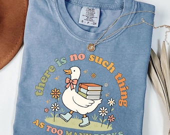 May include: A light blue Comfort Colors t-shirt with the phrase "there is no such thing as too many books." The graphic features a white duck wearing boots and carrying books, surrounded by flowers. A gold necklace is draped across the shirt.