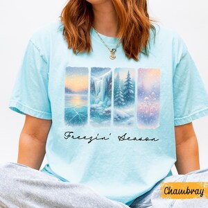 Puede incluir: Camiseta azul claro con un dise&ntilde;o de paisaje invernal. El dise&ntilde;o presenta cuatro paneles verticales que representan escenas nevadas, incluyendo un lago helado, una cascada y &aacute;rboles cubiertos de nieve. Las palabras "Freezin' Season" est&aacute;n escritas en cursiva debajo.