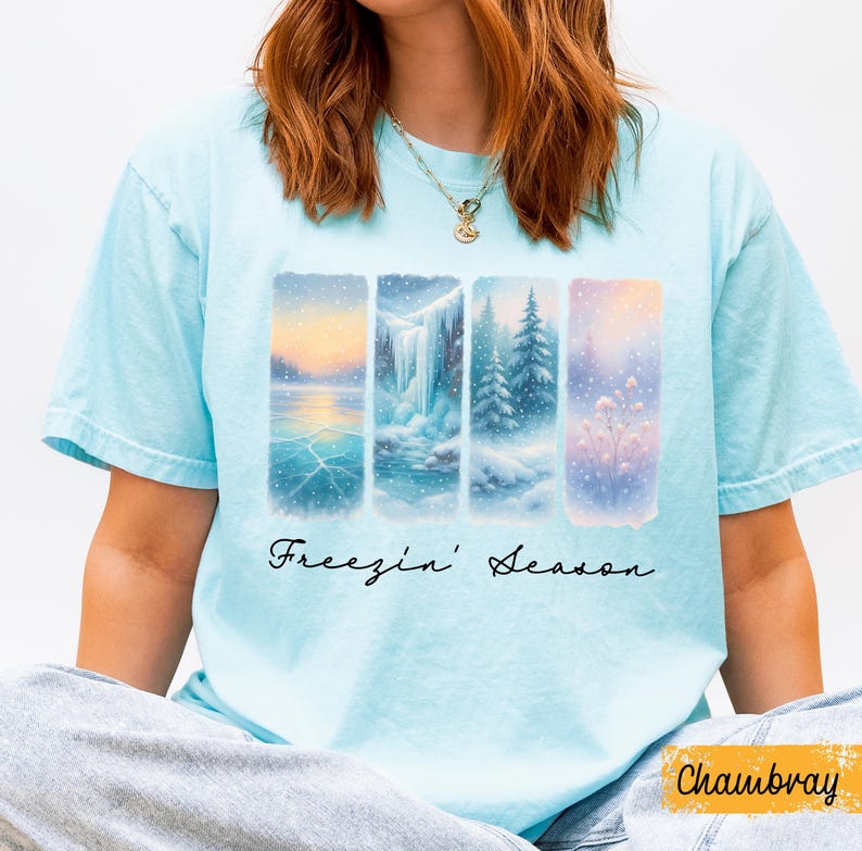 Puede incluir: Camiseta azul claro con un dise&ntilde;o de paisaje invernal. El dise&ntilde;o presenta cuatro paneles verticales que representan escenas nevadas, incluyendo un lago helado, una cascada y &aacute;rboles cubiertos de nieve. Las palabras "Freezin' Season" est&aacute;n escritas en cursiva debajo.