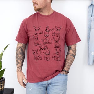 May include: A brick red short-sleeve t-shirt featuring a black line art graphic print of ten different Chihuahua dogs in various poses, including sitting, standing, and sleeping, arranged in a grid pattern across the center of the chest.