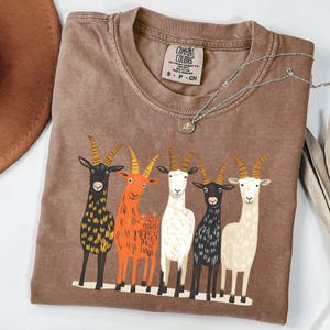 May include: Brown Comfort Colors t-shirt featuring a graphic of five colorful goats. The goats are in various colors, including black, orange, white, and cream. The shirt has a crew neck and short sleeves. A silver necklace with a pendant is on the shirt.