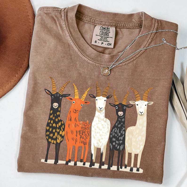 May include: Brown Comfort Colors t-shirt featuring a graphic of five colorful goats. The goats are in various colors, including black, orange, white, and cream. The shirt has a crew neck and short sleeves. A silver necklace with a pendant is on the shirt.