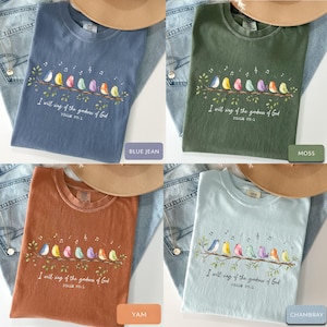 May include: A collection of four crew neck t-shirts in blue jean, moss, yam, and chambray colors. Each shirt features a graphic of eight colorful birds perched on a branch with musical notes and the text I will sing of the goodness of God Psalm 89:1.