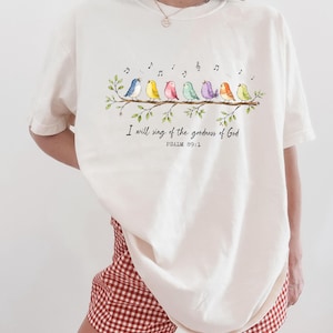 May include: An ivory t-shirt featuring a graphic of seven colorful birds perched on a leafy branch with musical notes above them. Below the branch, the text reads: I will sing of the goodness of God, PSALM 89:1. The shirt has a crew neckline.