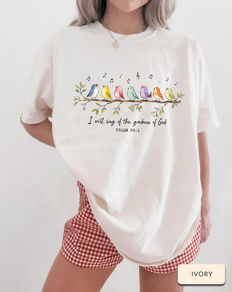 May include: An ivory t-shirt featuring a graphic of seven colorful birds perched on a leafy branch with musical notes above them. Below the branch, the text reads: I will sing of the goodness of God, PSALM 89:1. The shirt has a crew neckline.