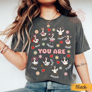 May include: Dark grey t-shirt with cartoon geese and positive words such as "Brave," "Kind," and "Unique." The words "YOU ARE" are in the centre. The design includes hearts and flowers.