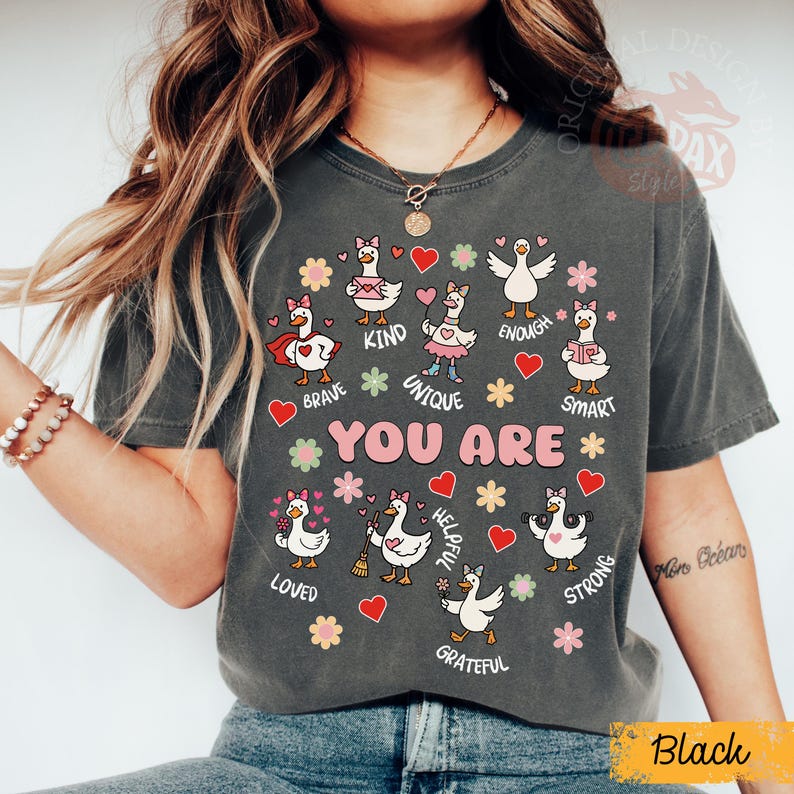 May include: Dark grey t-shirt with cartoon geese and positive words such as "Brave," "Kind," and "Unique." The words "YOU ARE" are in the centre. The design includes hearts and flowers.