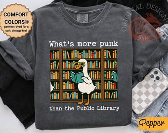 Puede incluir: Camiseta gris oscuro de manga larga con un diseño de un ganso con gafas leyendo frente a una estantería. El texto dice: "What's more punk than the Public Library". La camiseta tiene un tacto vintage.