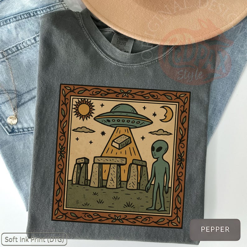 Op de afbeelding: Pepperkleurig t-shirt met een ingelijste afbeelding van een alien, Stonehenge en een UFO. Het ontwerp bevat een zon, maan en sterren. Het shirt wordt gecombineerd met een blauwe jeans en een bruine hoed. De tekst "Soft Ink Print (DTG)" is zichtbaar.