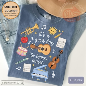 Op de afbeelding: Een blauw t-shirt met een grafische print met de tekst 'it's a good day to learn music' omringd door illustraties van een trompet, een lachende appel, een trommel, een fluit, een gitaar, een viool, een piano, een zon, een potlood en muzieknoten.
