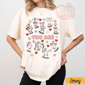 May include: An ivory-coloured t-shirt with a playful design. It features cartoon geese and the words "YOU ARE" in pink. The design includes words like "Brave," "Kind," and "Unique," along with hearts and flowers. The shirt has a relaxed fit.