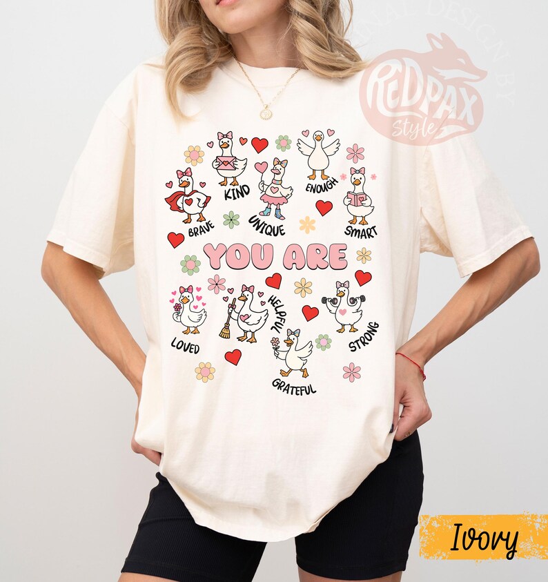 May include: An ivory-coloured t-shirt with a playful design. It features cartoon geese and the words "YOU ARE" in pink. The design includes words like "Brave," "Kind," and "Unique," along with hearts and flowers. The shirt has a relaxed fit.