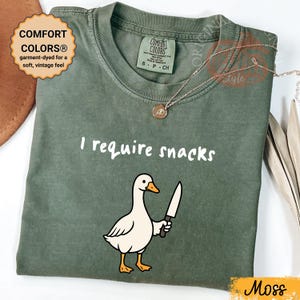 Puede incluir: Camiseta verde musgo con la frase "I require snacks" y un dibujo de un ganso con un cuchillo. La camiseta está teñida con la técnica garment-dye para un tacto suave y vintage. Un collar dorado con un colgante está sobre la camiseta.