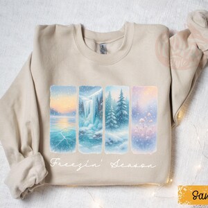 May include: Sand-colored sweatshirt featuring a winter landscape design. The design includes four rectangular panels depicting snowy scenes with a lake, waterfall, and trees. The text "Freezin' Season" is written in cursive below the panels.