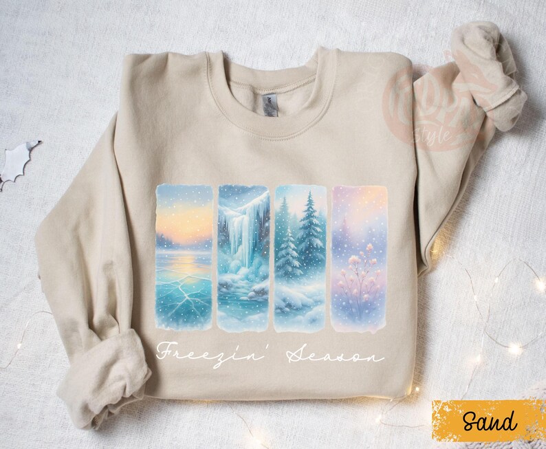 May include: Sand-colored sweatshirt featuring a winter landscape design. The design includes four rectangular panels depicting snowy scenes with a lake, waterfall, and trees. The text "Freezin' Season" is written in cursive below the panels.