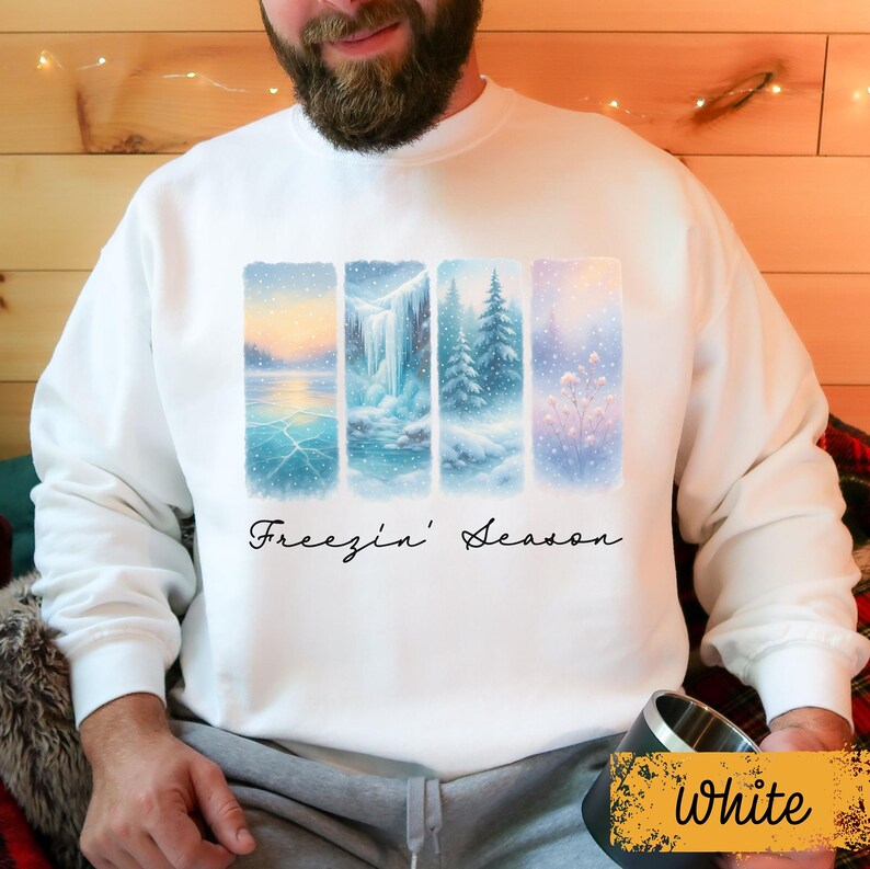 May include: White sweatshirt featuring a winter landscape design with four vertical panels. The panels depict snowy scenes with a lake, waterfall, evergreen trees, and a field of flowers. The text "Freezin' Season" is written in cursive.