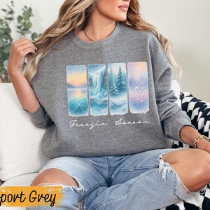 May include: Sport Grey sweatshirt featuring a winter landscape design. The graphic includes four rectangular panels depicting snowy scenes with a lake, waterfall, and trees. The words "Freezin' Season" are written in cursive below the panels.