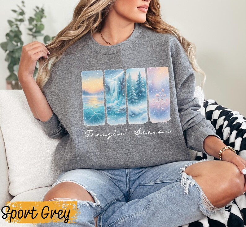 May include: Sport Grey sweatshirt featuring a winter landscape design. The graphic includes four rectangular panels depicting snowy scenes with a lake, waterfall, and trees. The words "Freezin' Season" are written in cursive below the panels.