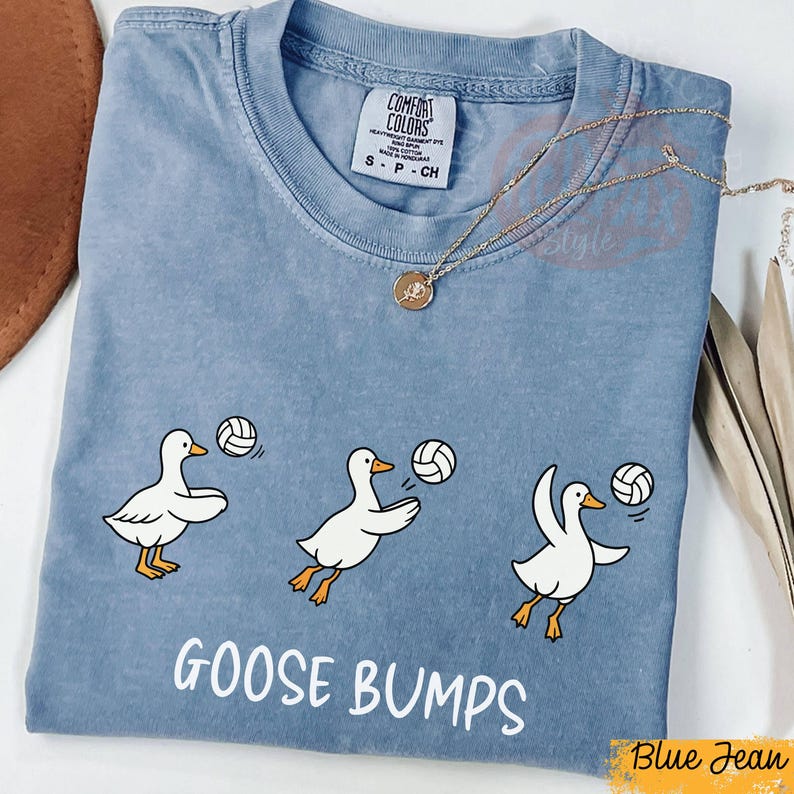 以下が含まれることがあります： 「GOOSE BUMPS」の白文字が入った青いTシャツ。3羽の白い漫画のガチョウがバレーボールをしているグラフィックが特徴です。シャツにはComfort Colorsのタグが付いています。