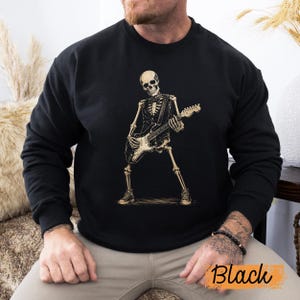 Peut inclure: Un sweat-shirt noir &agrave; manches longues avec une impression graphique repr&eacute;sentant un squelette portant un gilet et un n&oelig;ud papillon tout en jouant de la guitare &eacute;lectrique. Le squelette est beige clair et adopte une pose dynamique avec des baskets.