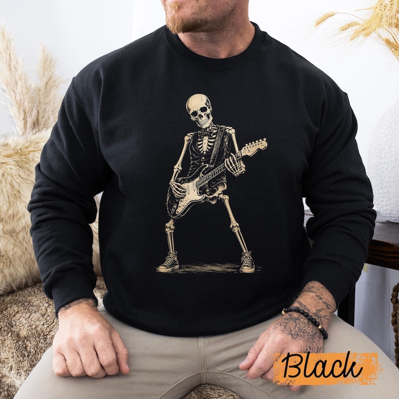 Peut inclure: Un sweat-shirt noir &agrave; manches longues avec une impression graphique repr&eacute;sentant un squelette portant un gilet et un n&oelig;ud papillon tout en jouant de la guitare &eacute;lectrique. Le squelette est beige clair et adopte une pose dynamique avec des baskets.