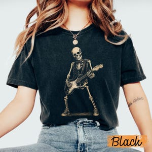 Peut inclure: Un t-shirt noir &agrave; manches courtes avec une impression graphique repr&eacute;sentant un squelette portant un gilet et un n&oelig;ud papillon tout en jouant de la guitare &eacute;lectrique. Le squelette est debout, porte des baskets, et le motif est en beige.