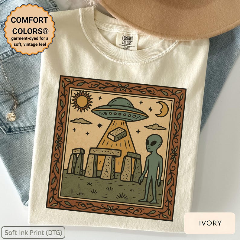Op de afbeelding: Ivoor kleurig T-shirt met een ingelijste afbeelding van een UFO, alien en Stonehenge. Het ontwerp bevat een zon, maan en sterren. Het shirt heeft de tekst "COMFORT COLORS&reg;" en "Soft Ink Print (DTG)".