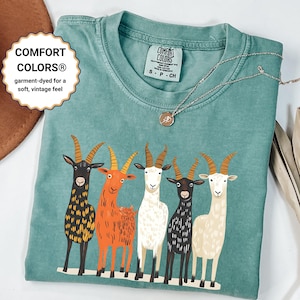 May include: A teal Comfort Colors t-shirt featuring a graphic of five stylized goats in various colors. The shirt has a crew neck and short sleeves. The text "COMFORT COLORS&reg; garment-dyed for a soft, vintage feel" is visible.