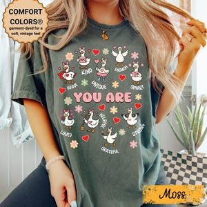 May include: A moss green Comfort Colors t-shirt featuring a graphic of geese and the words "YOU ARE" surrounded by positive affirmations. The shirt has a vintage feel and is garment-dyed for softness.