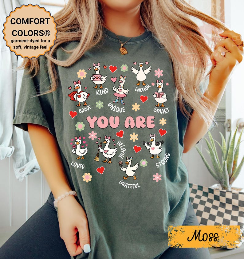 May include: A moss green Comfort Colors t-shirt featuring a graphic of geese and the words "YOU ARE" surrounded by positive affirmations. The shirt has a vintage feel and is garment-dyed for softness.