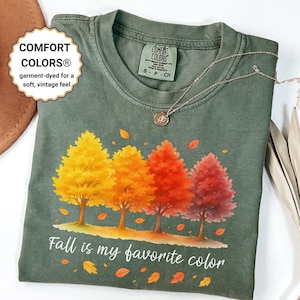 Puede incluir: Una camiseta de manga corta color verde oliva con un gráfico de cuatro árboles en colores otoñales amarillo, naranja, rojo y burdeos. El texto "Fall is my favorite color" aparece impreso bajo los árboles junto a hojas caídas.