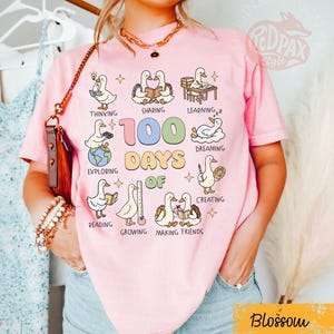 May include: Pink t-shirt with a graphic featuring cartoon ducks engaged in various activities like reading and creating. The text "100 Days of" is prominently displayed, along with the word "Blossom" in a yellow banner. The shirt is a casual style.