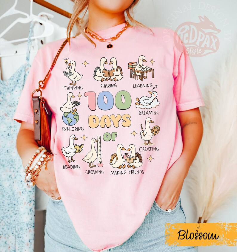 May include: Pink t-shirt with a graphic featuring cartoon ducks engaged in various activities like reading and creating. The text "100 Days of" is prominently displayed, along with the word "Blossom" in a yellow banner. The shirt is a casual style.
