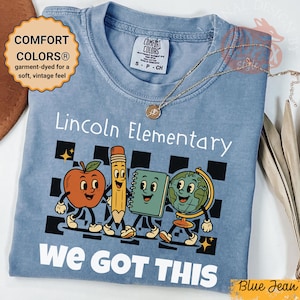 以下が含まれることがあります： 「Lincoln Elementary」と「We Got This」の白い文字が入った、ブルーのComfort ColorsのTシャツ。リンゴ、鉛筆、ノート、地球儀の漫画が描かれています。金のネックレスがシャツの上にかけられています。