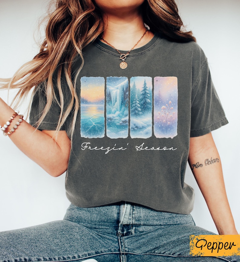 Puede incluir: Camiseta gris oscuro con un dise&ntilde;o de invierno. El dise&ntilde;o muestra cuatro paneles verticales con paisajes nevados, incluyendo un lago, una cascada y &aacute;rboles. Las palabras "Freezin' Season" est&aacute;n escritas en cursiva debajo.