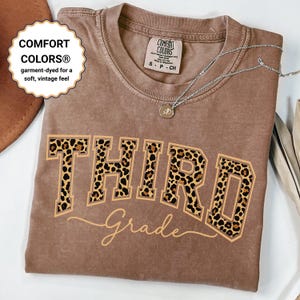 Könnte beinhalten: Ein braunes Comfort Colors T-Shirt mit dem Wort "THIRD" in Leopardenmuster-Buchstaben und "Grade" in Schreibschrift. Das Shirt hat ein weiches, vintage Gefühl. Eine silberne Halskette mit Anhänger befindet sich auf dem Shirt.