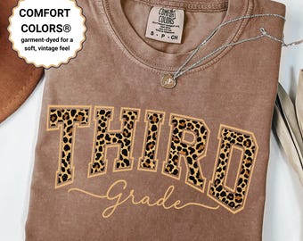 May include: A brown Comfort Colors t-shirt with the word "THIRD" in leopard print letters and "Grade" in cursive. The shirt has a soft, vintage feel. A silver necklace with a pendant is on the shirt.