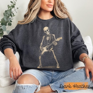 Peut inclure: Un sweat-shirt &agrave; col rond gris chin&eacute; fonc&eacute; avec une impression graphique beige repr&eacute;sentant un squelette jouant de la guitare &eacute;lectrique. Le squelette est repr&eacute;sent&eacute; debout, portant un gilet et des baskets, tenant le manche et le corps de la guitare.
