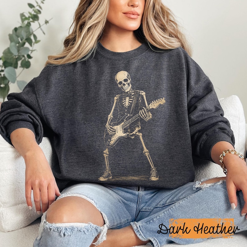 Peut inclure: Un sweat-shirt &agrave; col rond gris chin&eacute; fonc&eacute; avec une impression graphique beige repr&eacute;sentant un squelette jouant de la guitare &eacute;lectrique. Le squelette est repr&eacute;sent&eacute; debout, portant un gilet et des baskets, tenant le manche et le corps de la guitare.
