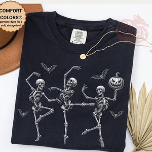 Puede incluir: Una camiseta negra con un gráfico de tres esqueletos blancos bailando con murciélagos y una calabaza de Halloween. La prenda cuenta con una etiqueta de la marca Comfort Colors en el cuello, escote redondo y mangas cortas con diseño impreso.