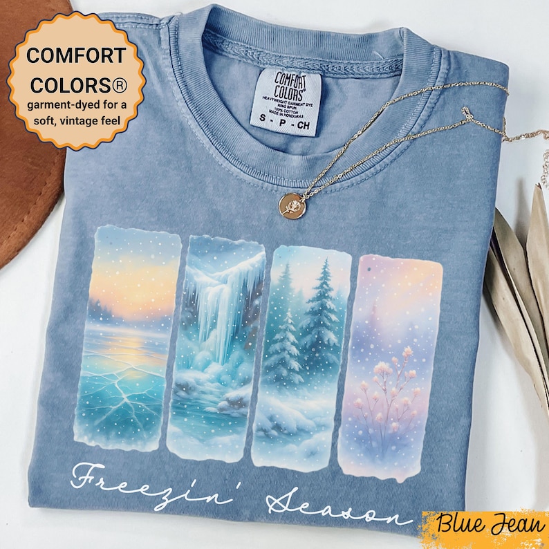 Puede incluir: Camiseta Comfort Colors azul vaquero con un gr&aacute;fico de escena invernal. El gr&aacute;fico presenta cuatro paneles rectangulares que representan un paisaje nevado con el texto "Freezin' Season". Un collar de oro con un colgante descansa sobre la camiseta.