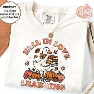 May include: Cream-colored t-shirt with a graphic featuring a duck holding books, pumpkins, and fall leaves. The text "FALL IN LOVE with LEARNING" is arched above the duck. The shirt has a vintage feel.