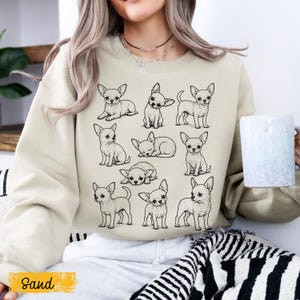 May include: A sand-colored crewneck sweatshirt featuring a graphic print of nine black and white line art illustrations of Chihuahua dogs in various poses, including sitting, standing, and lying down, arranged in a grid pattern on the front.
