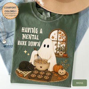 May include: A moss green t-shirt featuring a graphic of a ghost holding a whisk over a mixing bowl, a black cat, pumpkins, and a window with autumn leaves. The text reads "HAVING A MENTAL BAKE DOWN". The shirt has a Comfort Colors tag at the neck.
