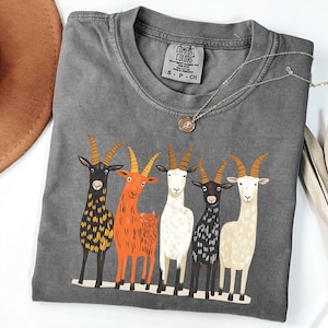 May include: A gray Comfort Colors t-shirt featuring a graphic of five colorful goats. The goats are in various colors, including black, orange, and white, with detailed horns. A silver necklace with a pendant is also visible.