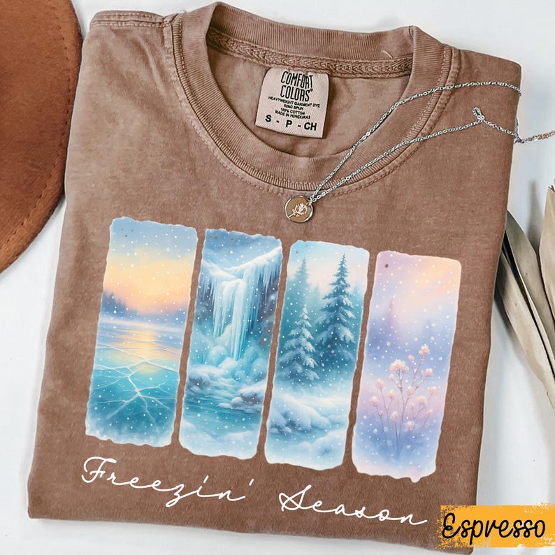 Puede incluir: Camiseta color espresso con un dise&ntilde;o de paisaje invernal y el texto "Freezin' Season". El dise&ntilde;o incluye cuatro paneles rectangulares que representan escenas nevadas. Un collar plateado con un colgante descansa sobre la camiseta.