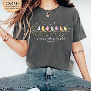 May include: A charcoal gray short-sleeve t-shirt featuring a graphic of seven colorful birds perched on a branch with musical notes above them. The text reads, "I will sing of the goodness of God" and "PSALM 89:1" in white lettering below the birds.