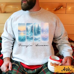 May include: A light gray sweatshirt featuring a winter landscape design with four vertical panels. The panels depict snowy scenes with a lake, waterfall, and trees. The text "Freezin' Season" is written in a cursive font below the panels.