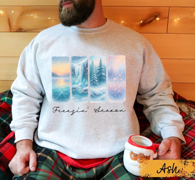 May include: A light gray sweatshirt featuring a winter landscape design with four vertical panels. The panels depict snowy scenes with a lake, waterfall, and trees. The text "Freezin' Season" is written in a cursive font below the panels.